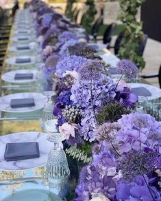 a long table is set with purple flowers and place settings for the guests to sit down