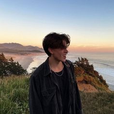 a young man standing on top of a lush green hillside next to the ocean at sunset