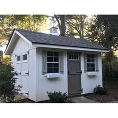 a small white shed with windows and shutters on the side, in front of some trees