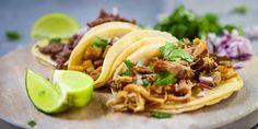 three tacos with meat, onions and cilantro on a cutting board next to lime wedges