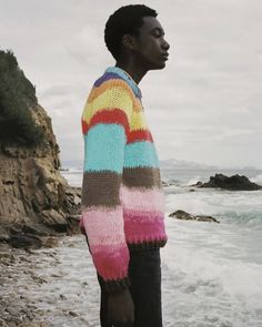 a man standing on top of a beach next to the ocean wearing a multicolored sweater