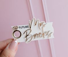 a person holding up a white and gold future mrs bookie sticker on pink background