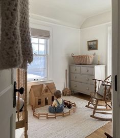 a child sitting on the floor in front of a rocking chair with a toy house next to it