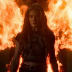 a woman with long hair standing in front of a large fire filled wall and flames behind her