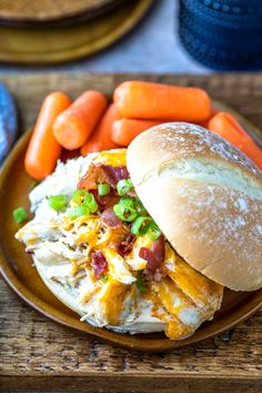 a chicken sandwich on a plate with carrots and potatoes next to it, sitting on a wooden table