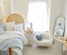 a bedroom decorated in white and blue with lights on the window sill, bed, chair, desk and mirror