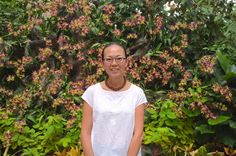 a woman wearing glasses standing in front of some plants and trees with purple flowers behind her