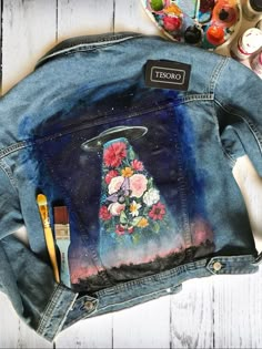 a jean jacket painted with flowers and an angel on it, next to paintbrushes