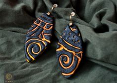 two earrings with designs on them sitting on a green fabric covered bed sheet, one is orange and the other is blue