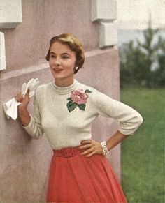 a woman in a white sweater and red skirt standing next to a building with her hands on her hips