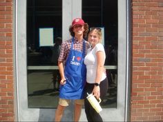 two people standing in front of a building with their arms around each other and one person wearing an apron