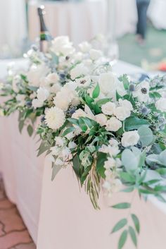 white flowers and greenery line the back of a long table at a wedding reception