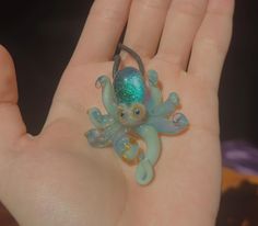 a hand is holding an octopus brooch in it's left hand, with the green and blue glass bead on top