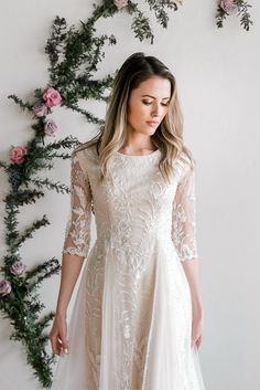 a woman wearing a white dress standing in front of a wall with flowers and greenery