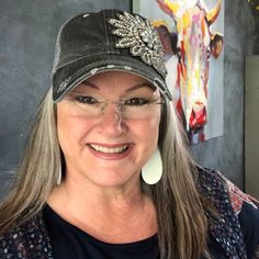 a woman with glasses and a hat smiling at the camera while standing in front of a cow painting