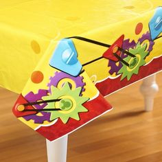 a yellow table cloth with colorful gears on it sitting on a wooden floor next to a white stool