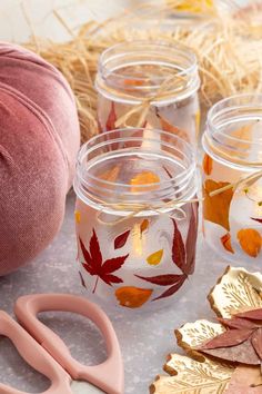 some glass jars with fall leaves on them next to pink scissors and other crafting supplies