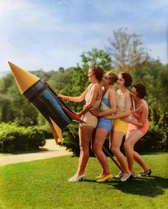 four women in bathing suits are holding a model rocket on the lawn, and one woman has her arm around another woman's neck