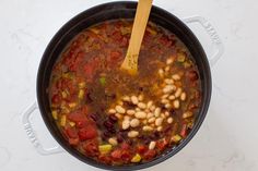 a pot filled with beans and vegetables on top of a white counter next to a wooden spoon