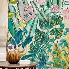 a table with a vase on top of it next to a wall covered in flowers and leaves