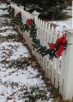 a white picket fence decorated with christmas lights and wreaths on top of it in the snow