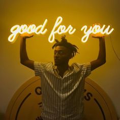 a man with dreadlocks standing in front of a neon sign that says good for you