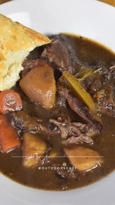 a white plate topped with meat and vegetable stew next to a piece of biscuit