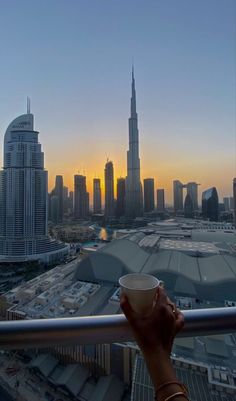 a person holding a coffee cup in front of a cityscape at sunset with the sun going down