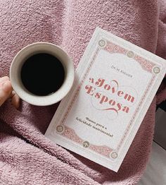 a person holding a cup of coffee next to a book on a pink bed spread