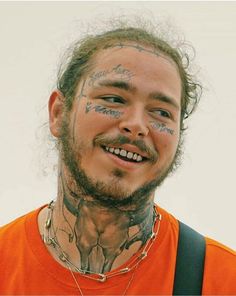 a man with long hair and tattoos on his face smiles at the camera while wearing an orange shirt