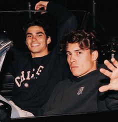 two young men sitting next to each other in a car, one holding his hand up