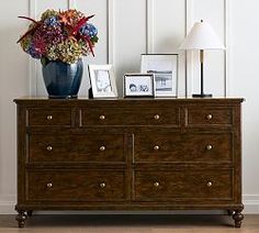 a wooden dresser with flowers and pictures on it next to a lamp in a room