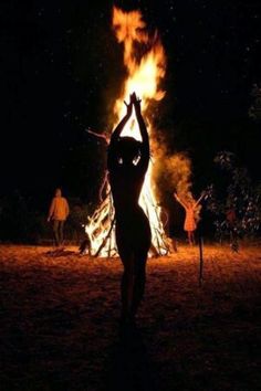 a woman standing in front of a fire with her arms up and hands raised above her head