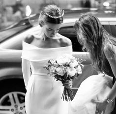 two women in white dresses standing next to each other near a car and one is holding a bouquet