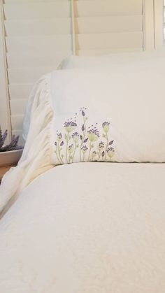 a bed with white linens and purple flowers on the pillowcase, along with a plant