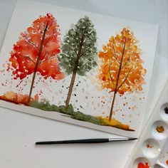 an artist's watercolor painting of three trees in fall colors with paintbrushes