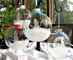 three glass ornaments with snow on them sitting on a white cloth covered tablecloth in front of a window