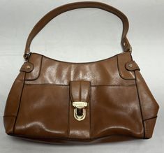 This Liz Claiborne handbag features a beautiful brown exterior with brass hardware. The bag has a spacious interior lined with polyester and includes a zip, button, and buckle closure. The top handle bag has a 20 inch shoulder strap and is perfect for any occasion, including travel, weddings, parties, casual outings, formal events, and business wear. The rectangular shaped bag measures 4inches in width, at it’s widest part the bottom . 8 inches in depth, and 8 inches in height. The exterior is made of faux leather with a gloss finish and is accented with brown vinyl top handle.The bag features one solid color pattern and is not vintage or handmade. There is a zippered middle partition taking up the width of the purse. 3 stitched in spacers in the lining opposite the front. A zippered pocke Brown Office Bag With Brass Hardware, Brown Satchel With Brass Hardware And Double Handle, Brown Bags With Brass Hardware And Double Handle, Classic Brown Bags With Metal Hardware, Brown Tote Shoulder Bag With Metal Hardware, Brown Satchel Bags With Brass Hardware, Brown Satchel With Brass Hardware For Office, Brown Satchel Shoulder Bag With Brass Hardware, Brown Flap Shoulder Bag With Metal Hardware