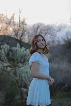 a woman in a white dress standing next to a cactus tree and looking at the camera