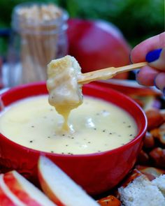 a person dipping cheese into an apple slice in a red bowl with apples around it