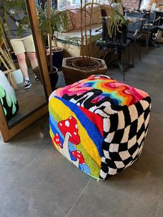 a multicolored foot stool sitting in front of a mirror on the floor next to a potted plant