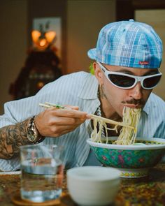 a man eating noodles with chopsticks in front of his face while wearing sunglasses and a hat