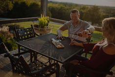 a man and woman sitting at a table on a deck with wine glasses in their hands