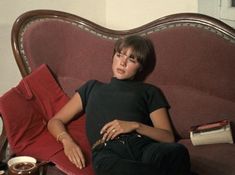 a woman laying on top of a red couch next to a cup of coffee and a book