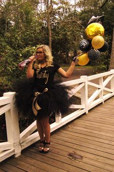 a woman in a black tutu holding balloons on a white bridge with trees and bushes behind her