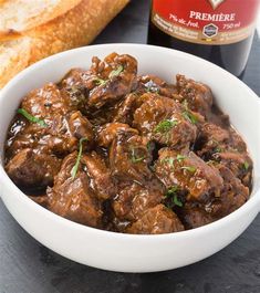 a white bowl filled with meat and bread next to a bottle of beer on a table