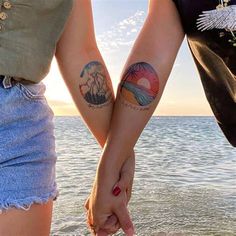 two people holding hands with tattoos on their arms and the ocean in the back ground