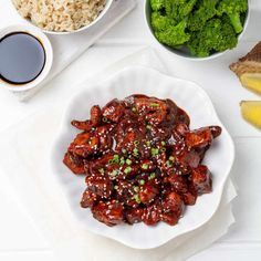 a white plate topped with meat covered in sauce next to bowls of rice and broccoli