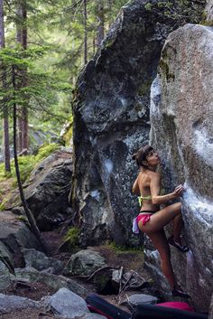 a woman climbing up the side of a large rock in a forest with backpacks