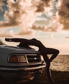 a woman leaning on the hood of a white car in front of water and clouds